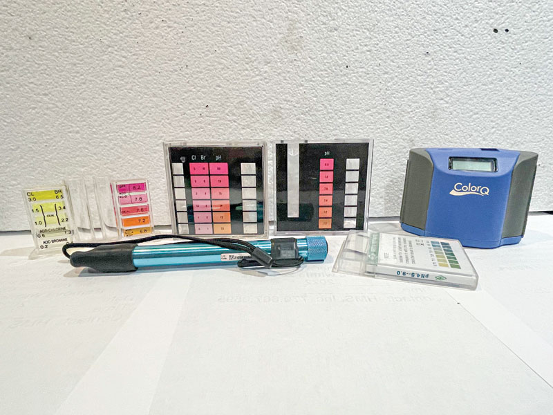 A collection of water testing equipment is displayed. Includes test strips, pH color charts, a blue digital meter, and a flashlight on a white surface.