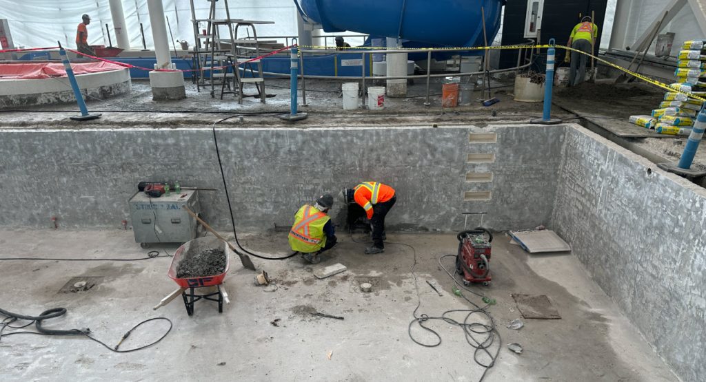 Workers in reflective vests repair a swimming pool at a construction site, surrounded by tools and materials.