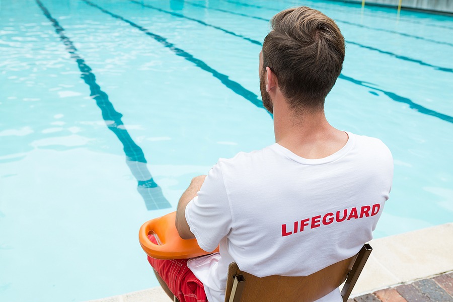 A lifeguard sits on a chair by a swimming pool, holding an orange buoy, wearing a white shirt with 