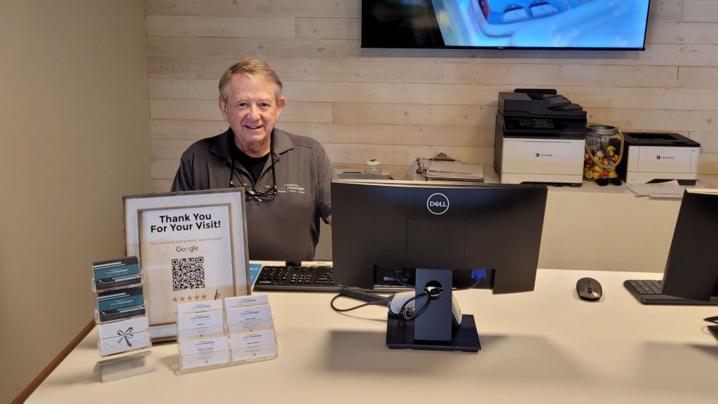 Man at a reception desk smiling, with a computer and a sign promoting Google reviews in a light wood-paneled office.