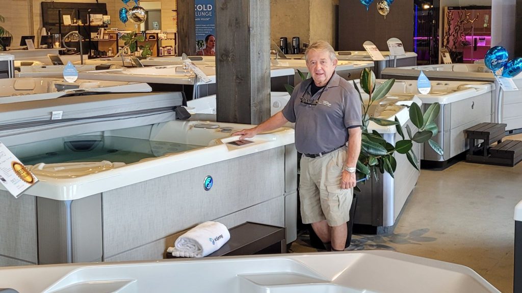 A middle-aged man stands beside a lit hot tub in a display showroom, surrounded by additional hot tubs and promotional materials, with blue balloons in the background.