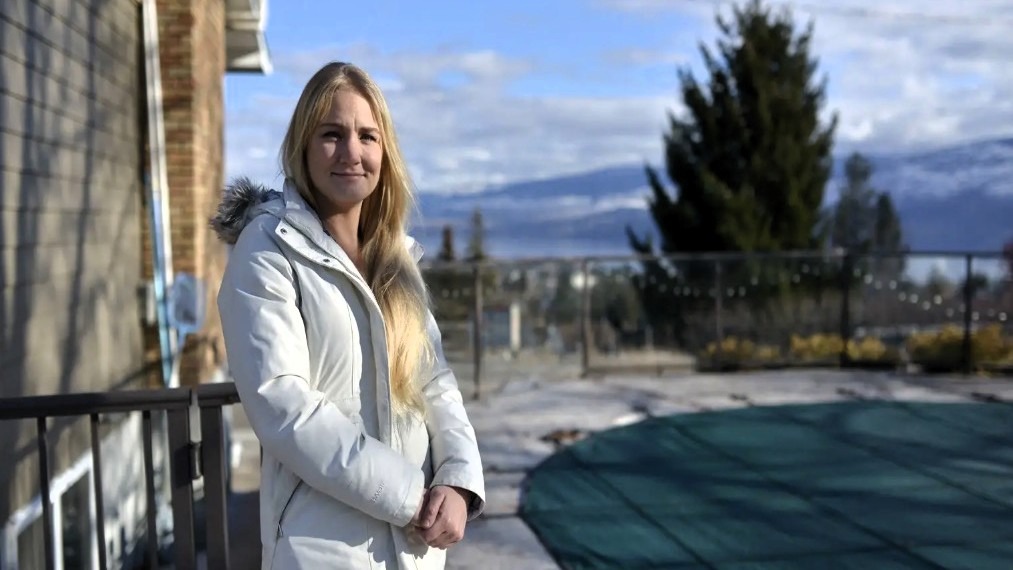 A young woman with long blonde hair stands on a balcony wearing a light-colored winter coat, with a green pool cover and mountains in the background.