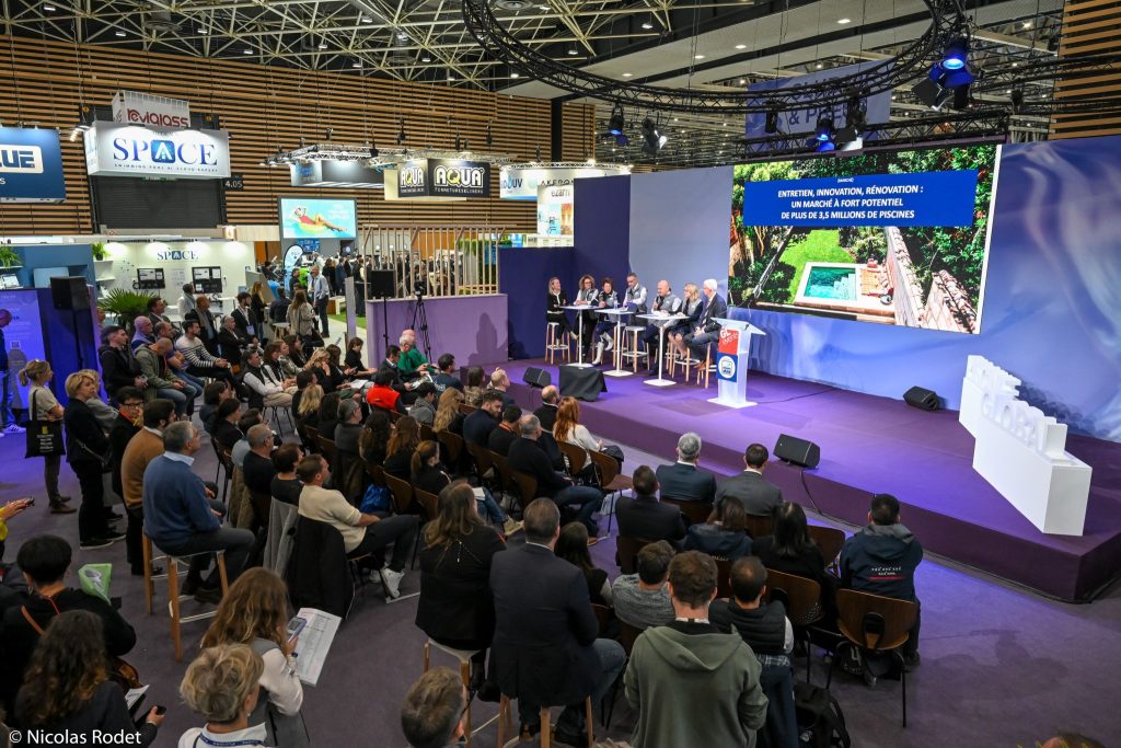 A conference presentation with a seated audience watching a panel of speakers on stage, featuring a screen displaying a swimming pool image. Various promotional banners are visible in the background.