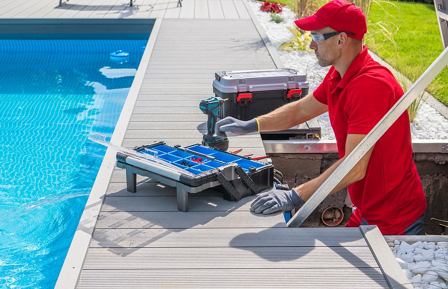 A male worker in a red shirt and cap repairs equipment near a blue swimming pool, utilizing a toolbox filled with tools on a wooden deck.