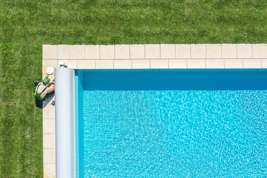 Aerial view of a swimming pool with clear blue water, surrounded by grass. A person is crouched near the pool performing maintenance tasks, with a white pool cover roller visible.
