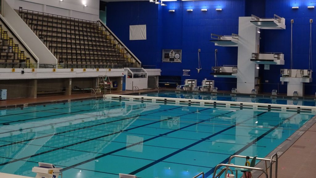 An indoor swimming pool with clear blue water, marked lanes, metal bleachers, and diving platforms against a blue wall.