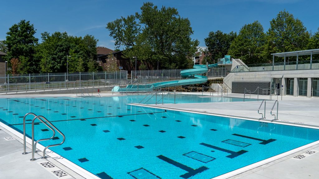 An outdoor swimming pool with clear blue water, a light turquoise water slide, and stainless steel ladders, surrounded by trees and a concrete deck.