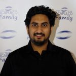 Portrait of a smiling bearded man in a black shirt standing in front of an “ACAPULCO Family” branded backdrop.