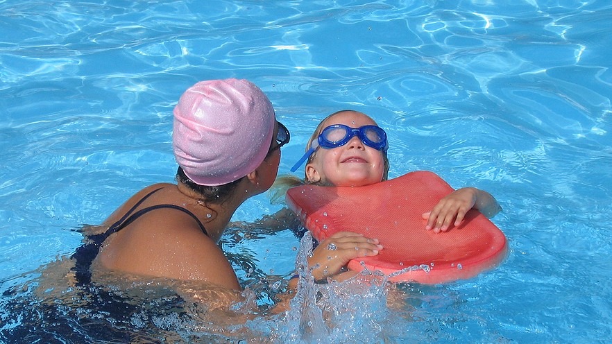 Adult swimmer assists a young swimmer wearing goggles and resting on a red flotation device in a sunlit pool.