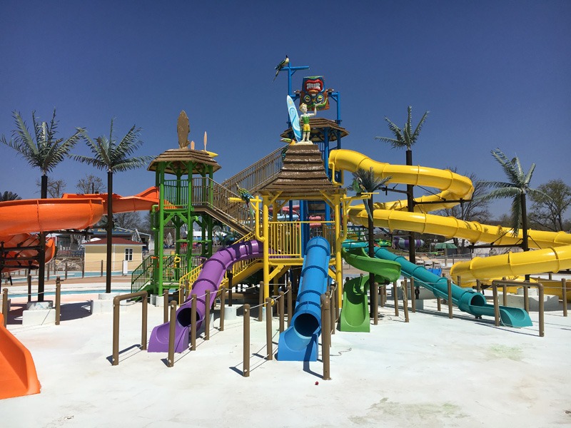 Colorful multi-level water-play structure with numerous orange, purple, blue, green, teal, and yellow slides surrounded by palm trees.