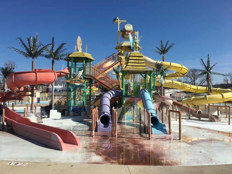 Colorful outdoor water-play structure with multiple twisting slides, palm-tree decorations, and wet concrete beneath a clear blue sky.
