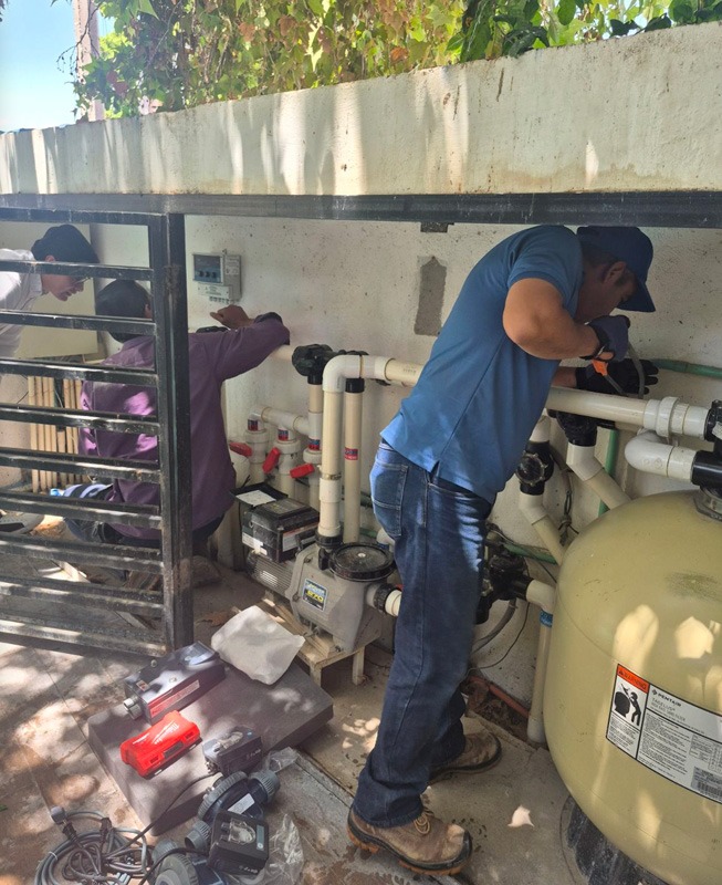 Two workers repair pool filtration equipment inside an outdoor utility enclosure.