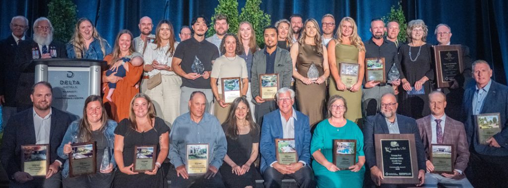 Large group posing with awards on a stage in front of blue curtains and greenery.