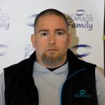 Portrait of a man wearing a black vest with blue trim in front of an Acapulco Family promotional backdrop.