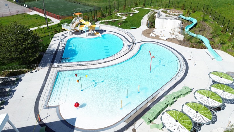 Two distinct pool areas Aerial view of an outdoor aquatic center with two turquoise pools, colorful water-play features, a winding blue water slide, and patterned shade umbrellas.