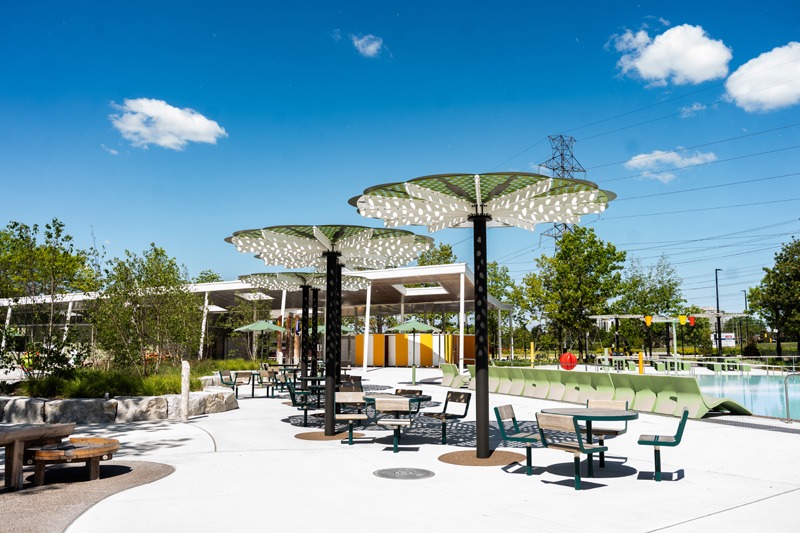 Panoramic view of an outdoor aquatic center with pools, yellow-and-white water slides, landscaped walkways, and surrounding trees.