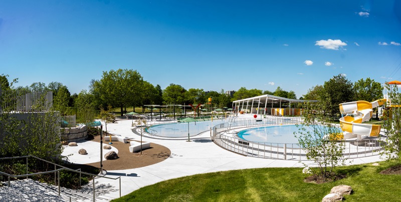 A transformed facility Panoramic view of an outdoor aquatic center with pools, yellow-and-white water slides, landscaped walkways, and surrounding trees.