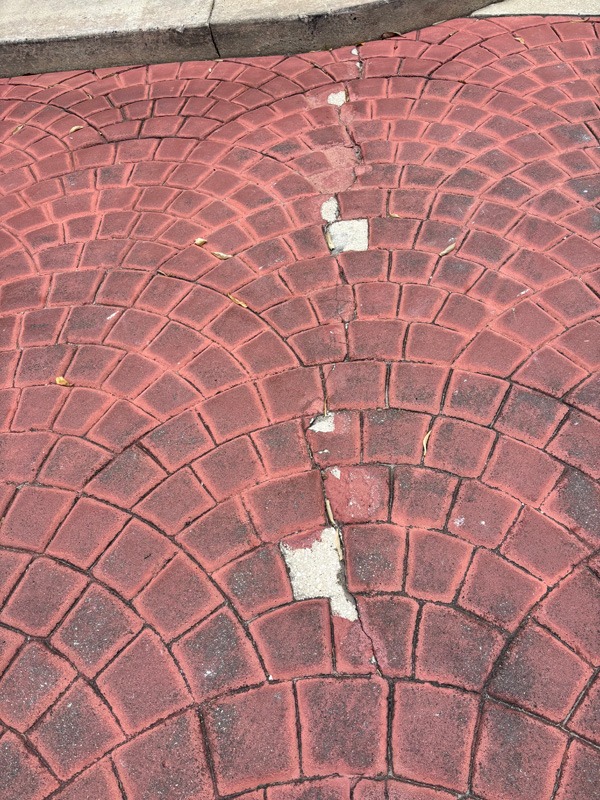 Close-up of weathered red brick paving in a curved fan pattern, with a central crack, white repair patches, and a concrete curb.