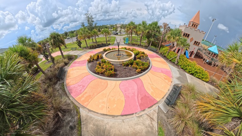 Colorful circular plaza with a central fountain, tropical landscaping, and a castle-like building beside the water.