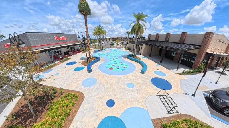 A sunny aerial view of a colorful shopping plaza with palm trees, storefronts, and a blue-and-turquoise splash pad decorated with geometric shapes.