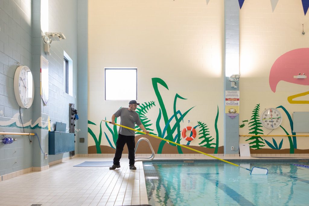 Person uses a long yellow pool skimmer beside an indoor swimming pool decorated with aquatic murals.