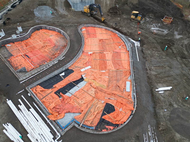 Kidstown construction Aerial view of a muddy construction site with two curved, orange-covered foundation areas, excavators, workers, and bundles of white pipes.