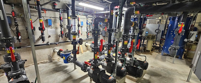 Mechanical utility room filled with interconnected pipes, valves, pumps, gauges, and a large blue filtration tank.