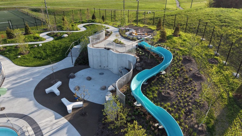 Aerial view of a landscaped playground with a large winding turquoise slide, concrete play structure, curved paths, and surrounding grassy fields.