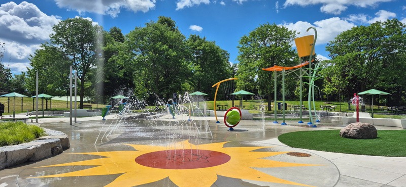 Colorful outdoor splash pad with spraying water jets, sunburst-painted pavement, shade umbrellas, and water-play structures surrounded by trees.