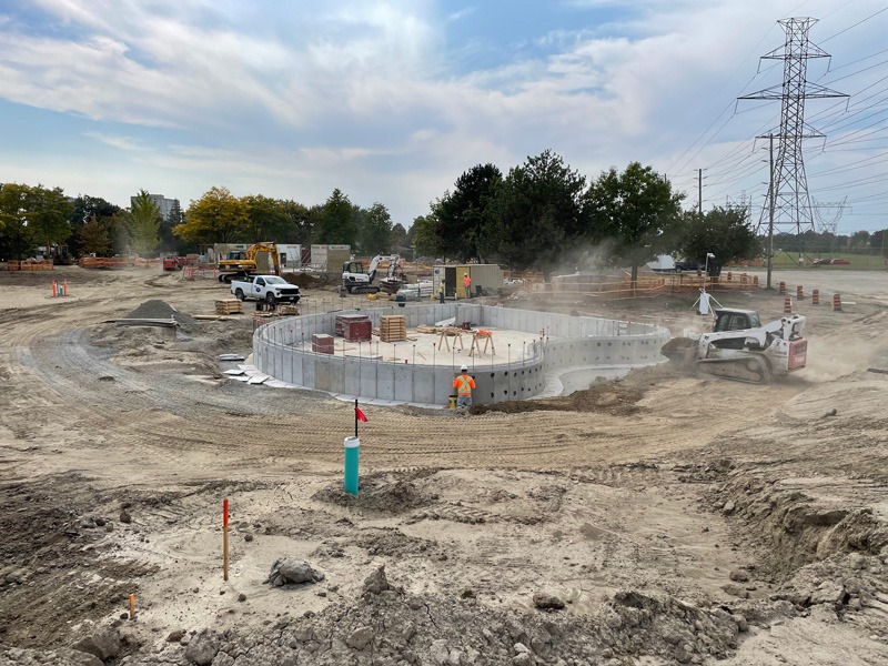 Reinforced casts Active construction site with a circular concrete structure, heavy equipment, workers, and a large electrical transmission tower in the background.