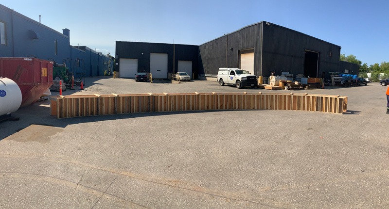 Curved formwork Industrial yard with a long wooden-slatted structure across the foreground and warehouses, vehicles, and equipment in the background.