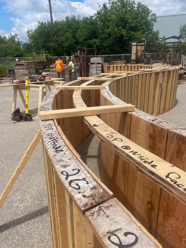 Curved wooden construction forms with handwritten markings fill an outdoor work area, with two high-visibility-vested workers and a yellow level in the background.