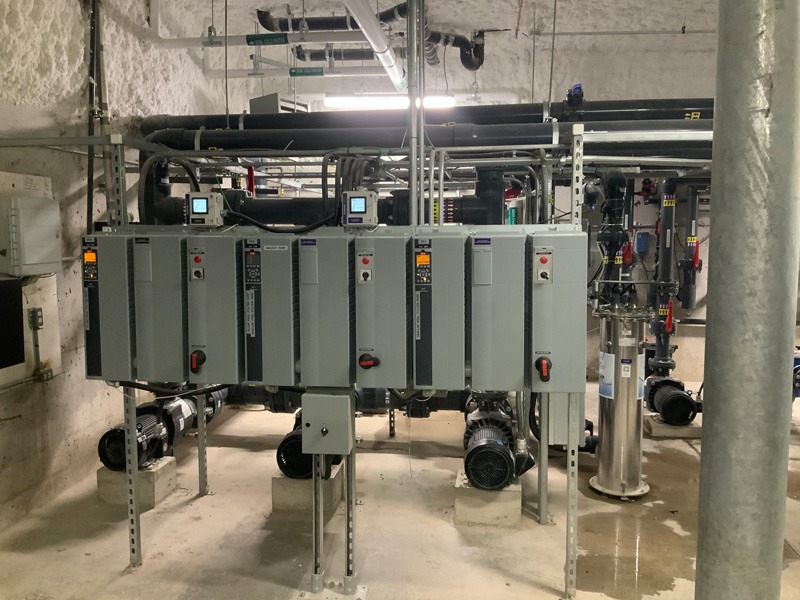 Industrial mechanical room with a row of gray electrical control cabinets, pumps, pipes, and other utility equipment.