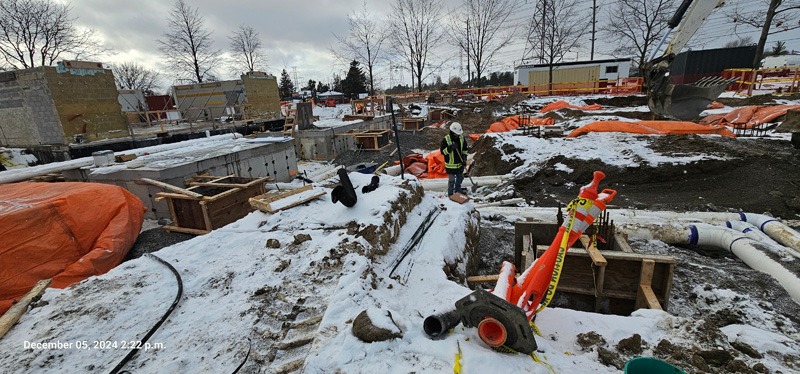 Winter construction Snow-covered construction site with a worker in safety gear standing beside an excavated trench.