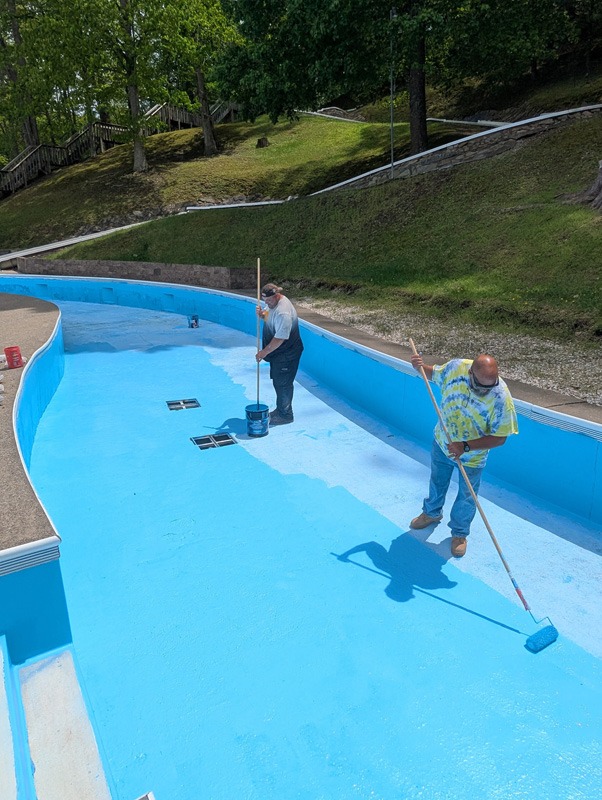 Two workers paint the bright-blue interior of an empty swimming pool with long-handled rollers.
