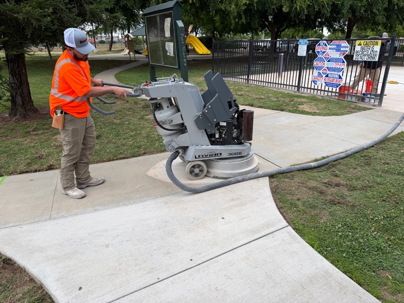 Worker operating a concrete floor grinder on a curved walkway beside a fenced playground.