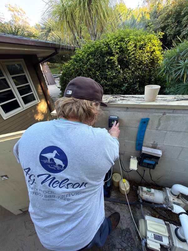 Person in a gray service shirt operates pool equipment beside a cinder-block wall surrounded by dense greenery.