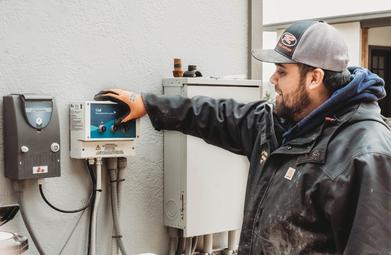 Worker in protective clothing operates a wall-mounted control panel connected to pipes and utility equipment.