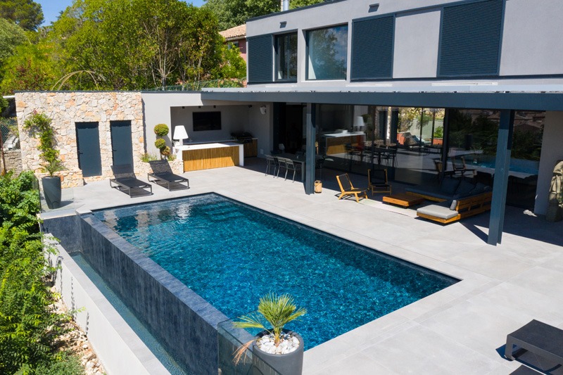 Modern two-story home with a vivid blue rectangular pool, stone wall, covered patio, and outdoor seating surrounded by greenery.