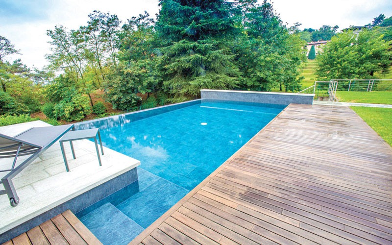 Blue backyard swimming pool bordered by a wooden deck, lounge chairs, and dense green trees.