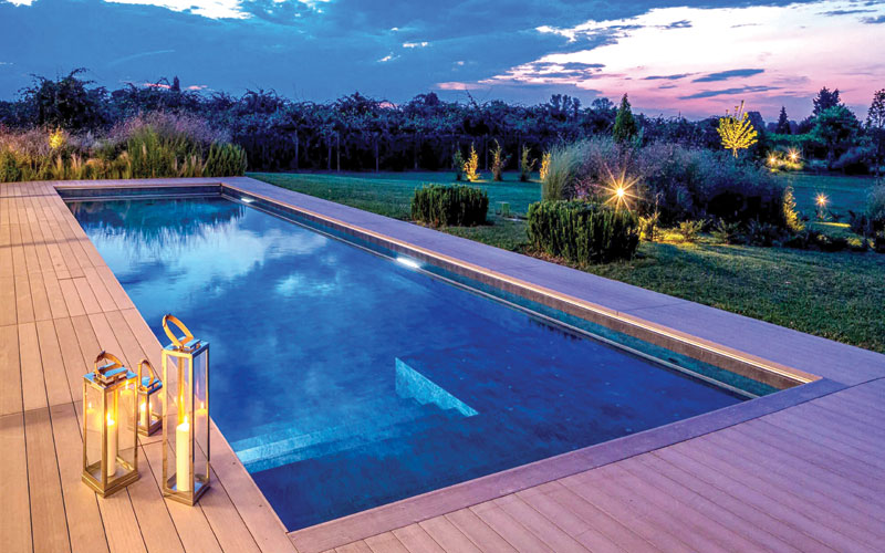 Rectangular illuminated swimming pool surrounded by a wooden deck and landscaped garden at dusk.