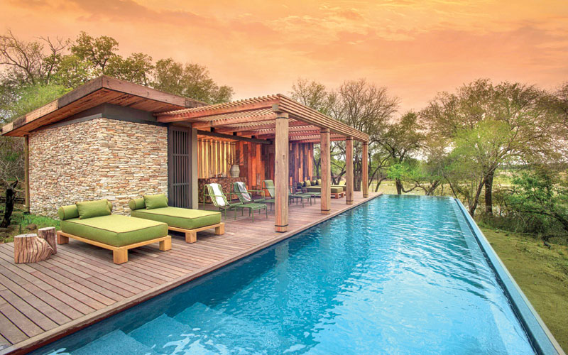 Modern wood-and-stone lodge with green lounge furniture beside a bright blue pool surrounded by trees at sunset.