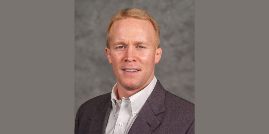 Head-and-shoulders portrait of a man with short reddish-blond hair wearing a dark blazer and light button-down shirt against a gray background.