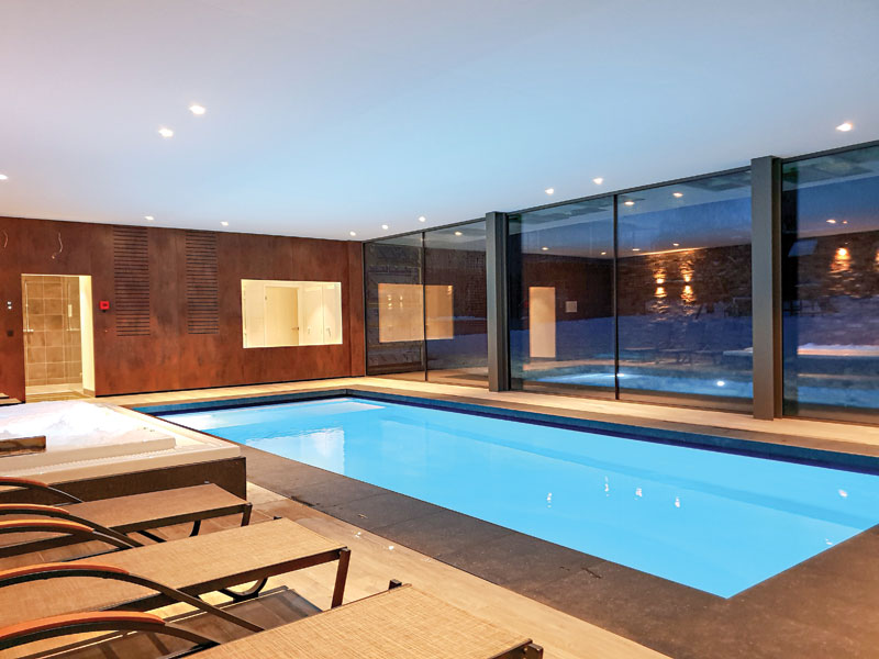 Modern indoor swimming pool with blue water, glass walls, and lounge chairs in the foreground.