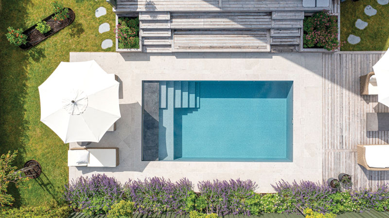 Overhead view of a turquoise rectangular pool surrounded by a pale patio, white umbrella, wooden decking, and lush garden landscaping.